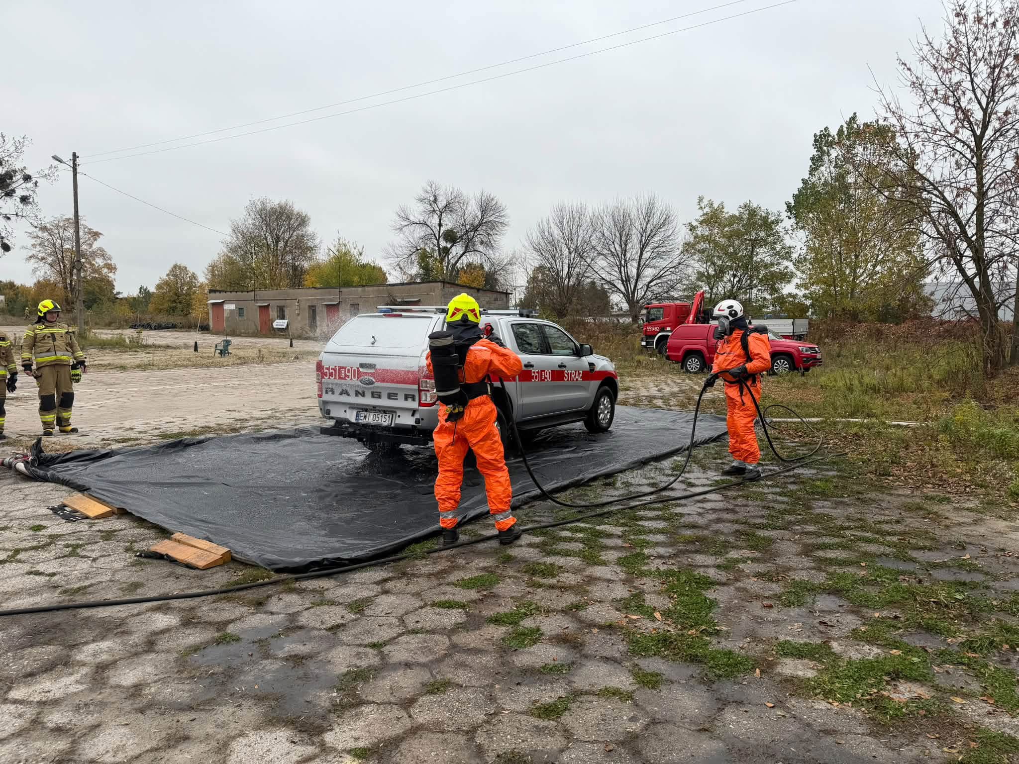 Strażacy w kombinezonach ochronnych przy stanowisku dekontaminacji pojazdu.