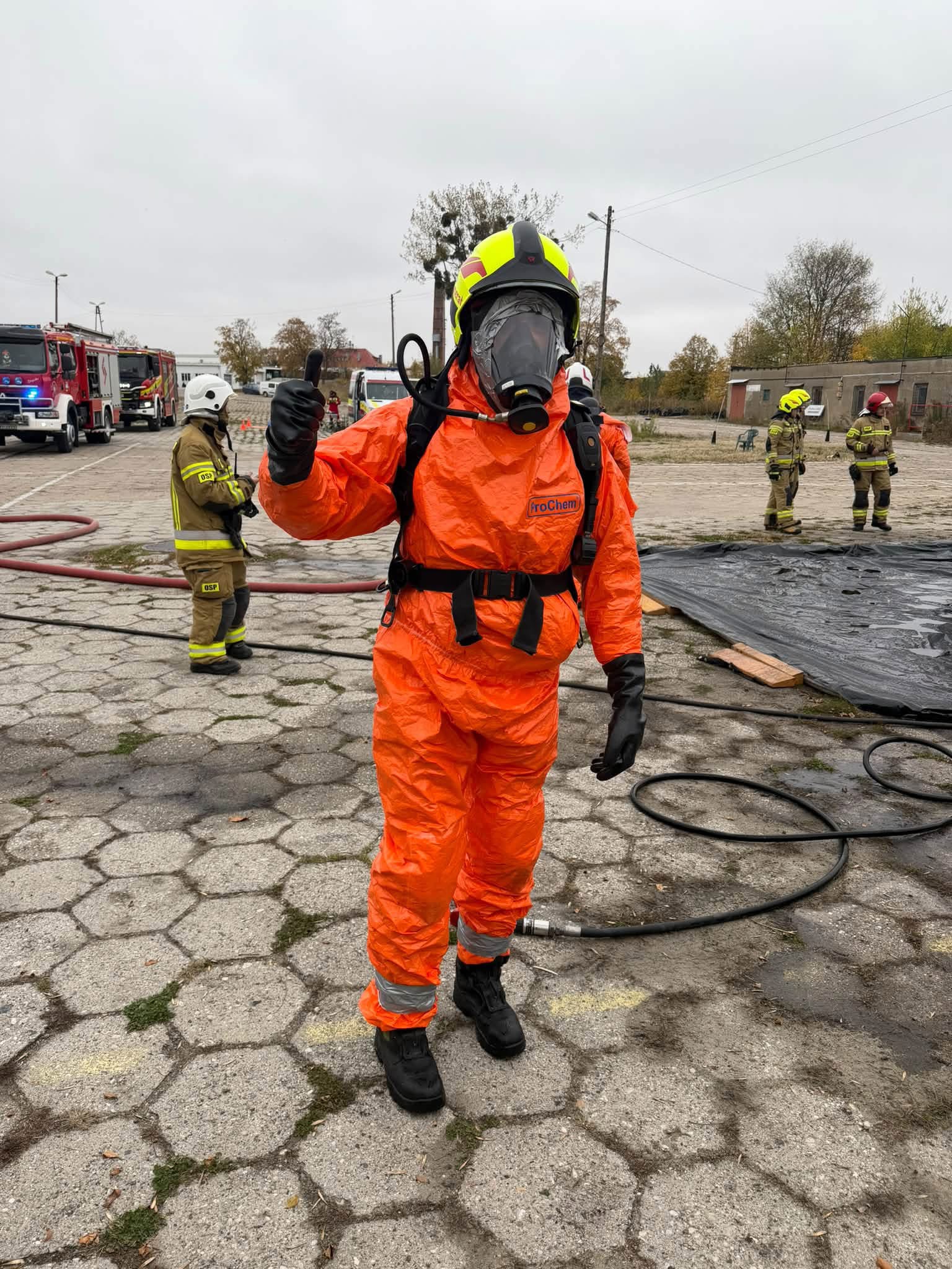Strażak w pomarańczowym kombinezonie i masce ochronnej pokazuje gest kciuka w górę.