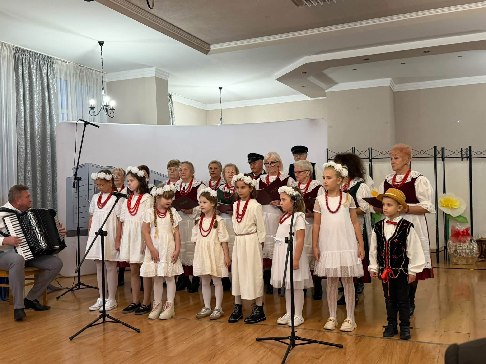 Uczniowie w białych strojach i koralach oraz członkowie zespołu w trakcie wspólnego śpiewania.