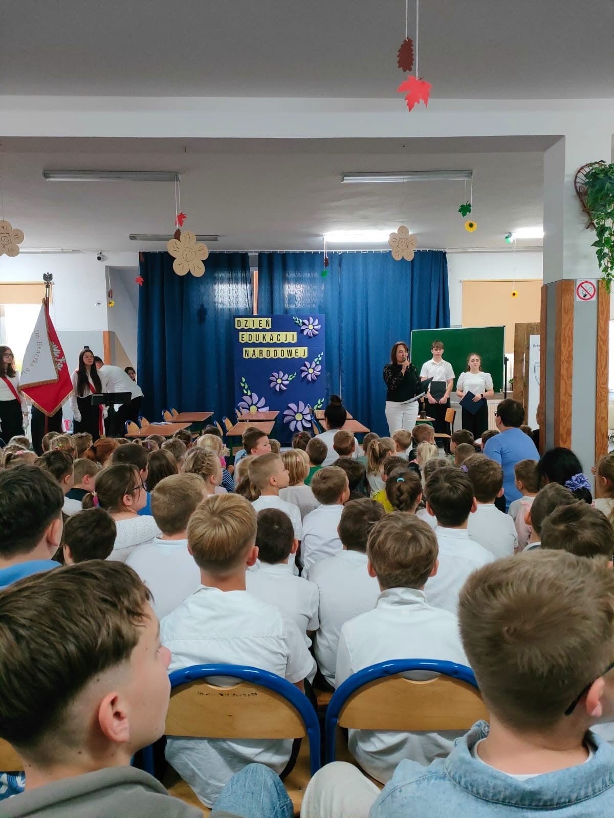 Scenografia szkolnego przedstawienia z napisem Dzień Edukacji Narodowej, dekorowana papierowymi kwiatami. Fot. Szkoła Podstawowa im. Henryka Sienkiewicza w Wierzchlesie.